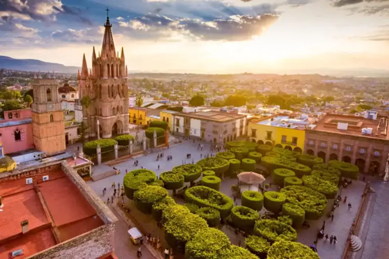 Aerial view of a historic town square at sunset, featuring a tall Gothic cathedral and neatly clipped topiary gardens.