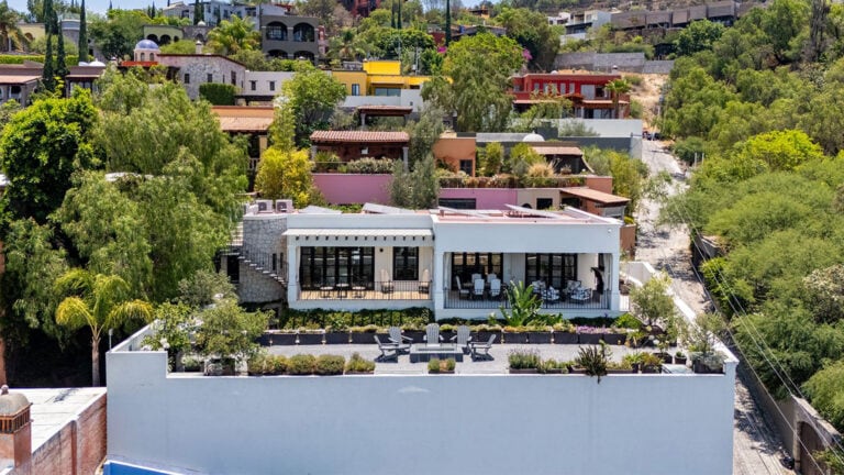 Hillside neighborhood with colorful houses and lush trees; in the foreground, a white modern villa with a covered patio and outdoor seating.