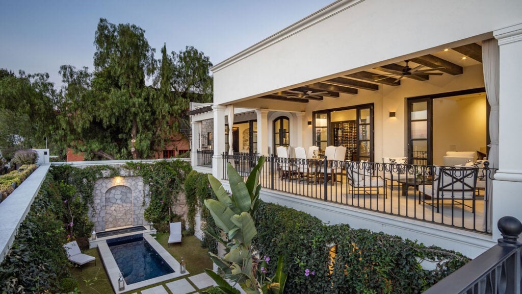 Wide view of a white Mediterranean-style house with a covered terrace, outdoor dining area, ceiling fans, and a long rectangular pool surrounded by greenery and stone walls.