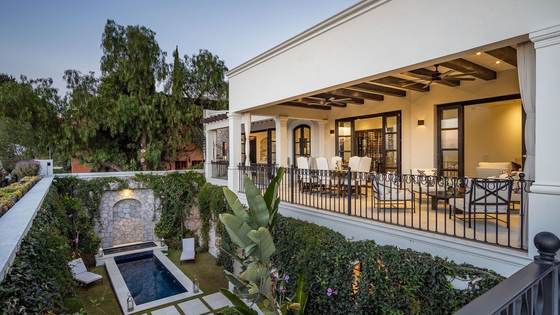 Wide view of a white Mediterranean-style house with a covered terrace, outdoor dining area, ceiling fans, and a long rectangular pool surrounded by greenery and stone walls.