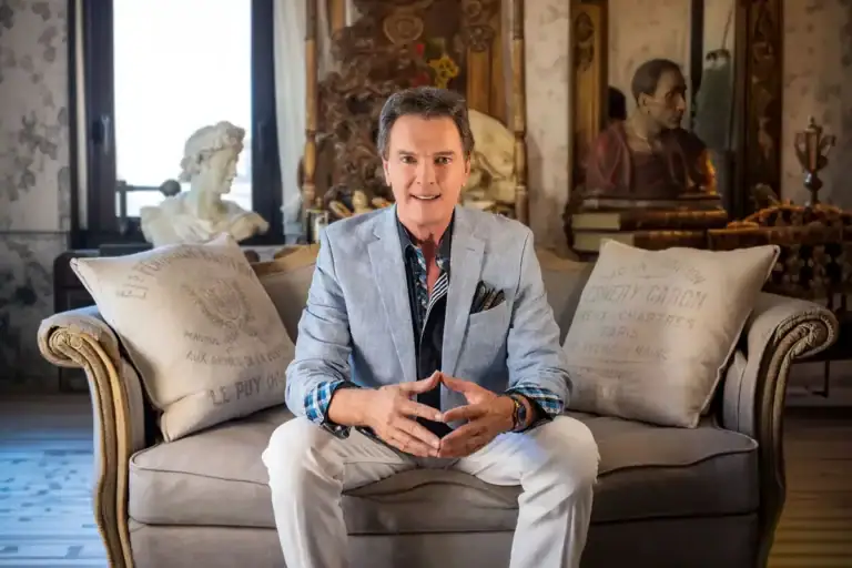 Man in a light gray blazer sits on a beige sofa, hands clasped, facing the camera in a vintage-filled living room.