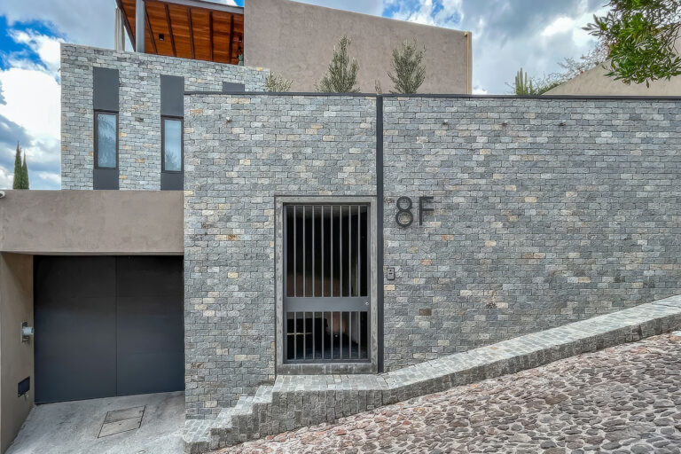 Gray stone building façade with a black metal gate and the number 8F on the wall; a cobblestone ramp/sidewalk leads to the entrance.