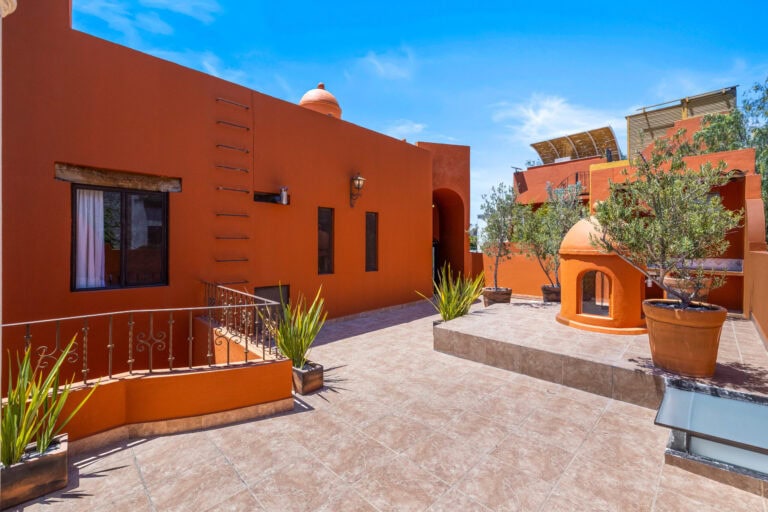 Orange adobe-style courtyard with arched doorway, potted plants, and decorative iron railing under a bright blue sky.