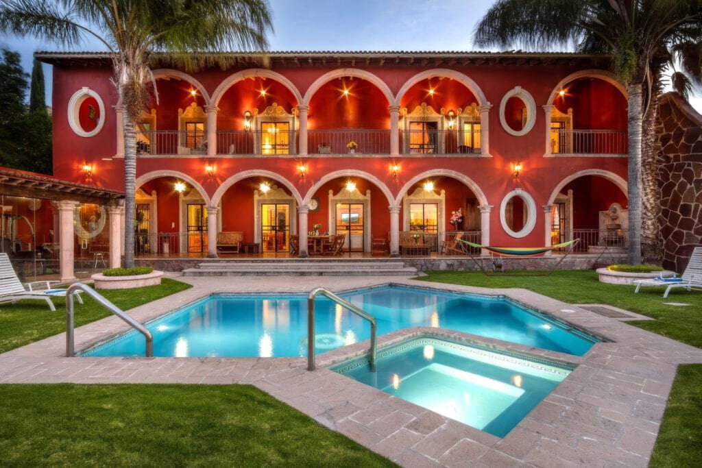 Red Mediterranean-style hotel building with white arches and balconies, palm trees, and a turquoise pool in foreground. (Informative)