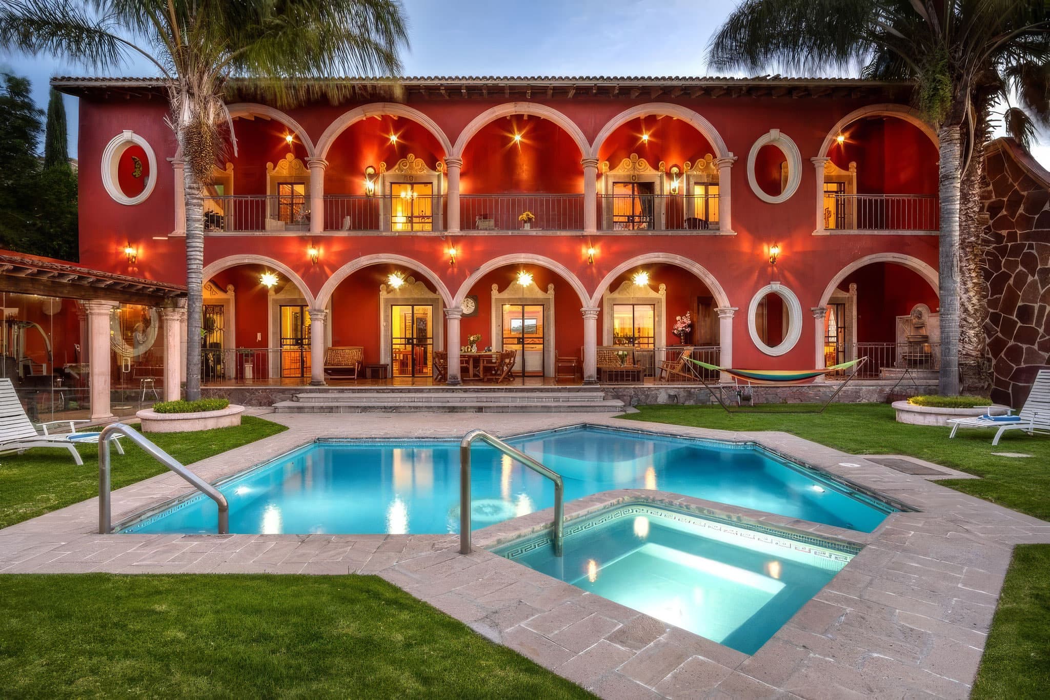 Red Mediterranean-style hotel building with white arches and balconies, palm trees, and a turquoise pool in foreground. (Informative)