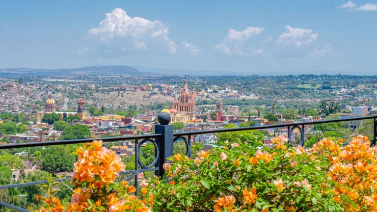 Cityscape with colorful buildings and church spires, seen from a balcony railing with orange flowers in the foreground.