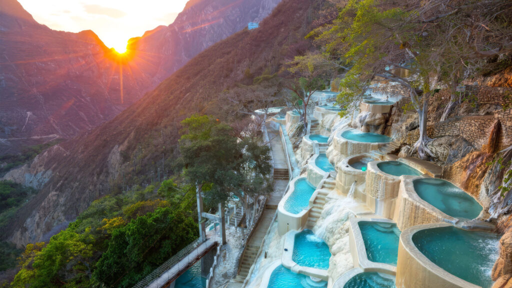 Sunset behind a mountain as turquoise terraced pools cascade down a rocky mountainside along a hillside path.