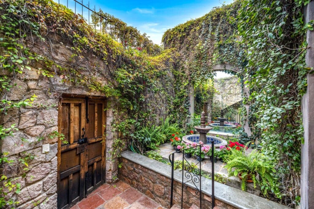 Stone-walled courtyard covered in ivy with a wooden door on the left and a circular fountain surrounded by potted flowers.