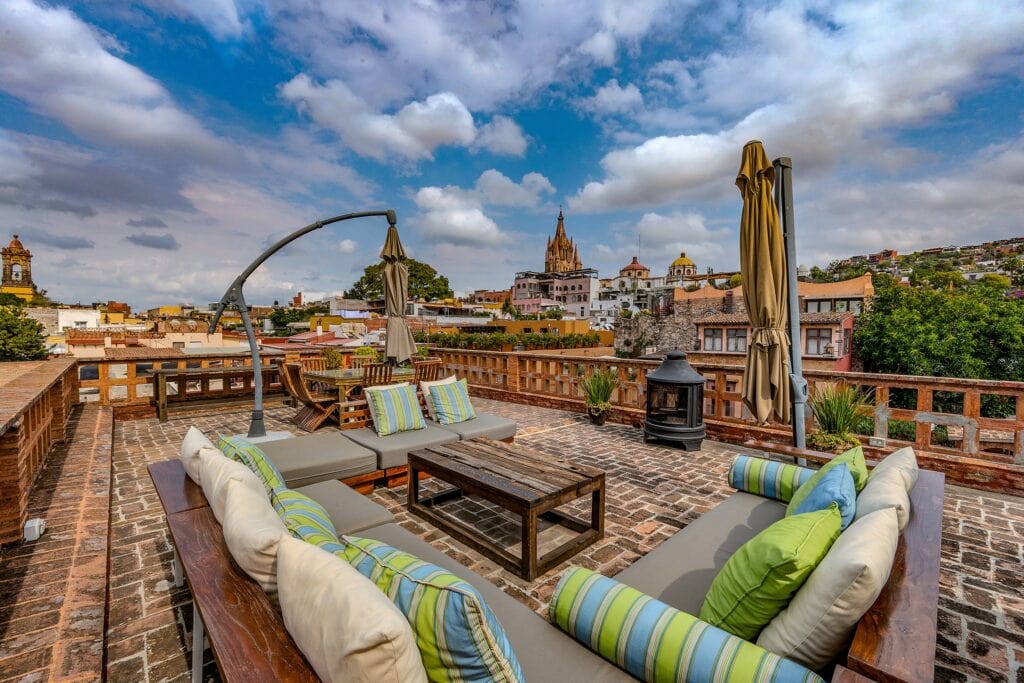 Rooftop terrace with cushioned outdoor sofas and pillows, overlooking a historic town with church spires under a blue, cloudy sky.