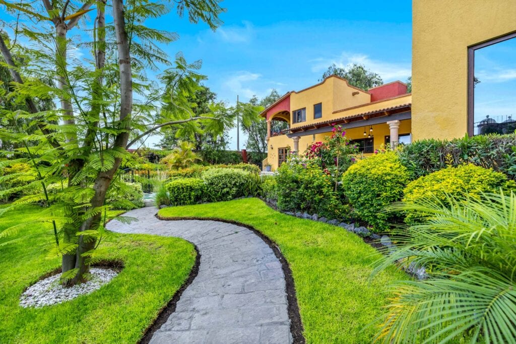 Winding stone path through a lush garden toward a yellow Mediterranean-style house with an arched porch.