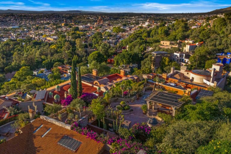 Panoramic view of a hillside town with red-tiled roofs, lush trees, and distant mountains under a bright blue sky.