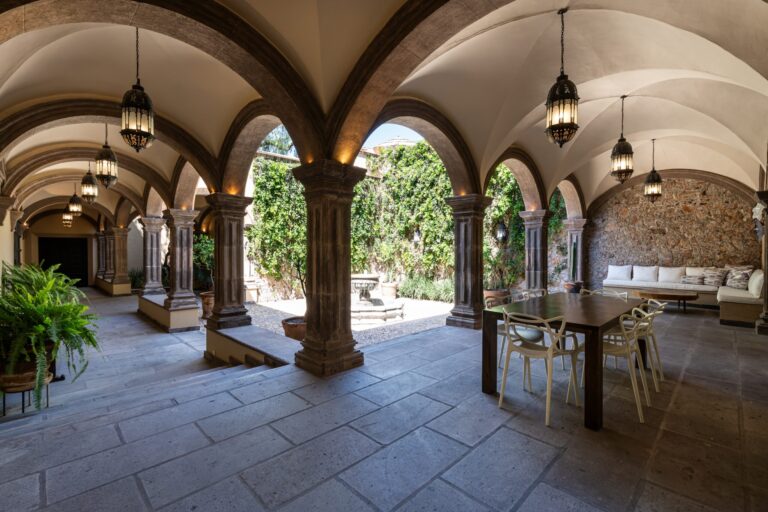 Sunlit open-air courtyard with stone arches, hanging lanterns, and a long wooden table with cream chairs along a colonnade, San Miguel de Allende