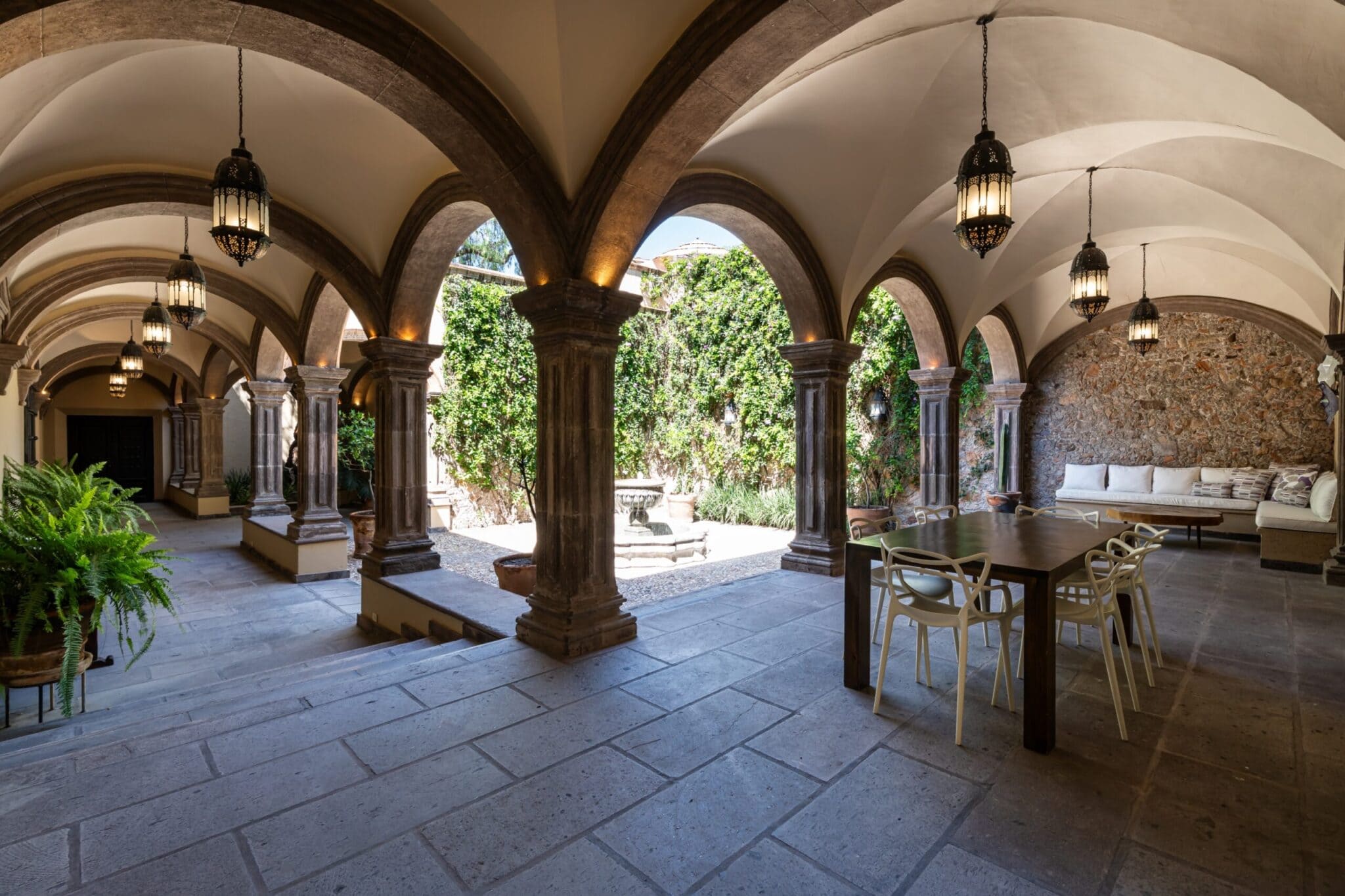 Sunlit open-air courtyard with stone arches, hanging lanterns, and a long wooden table with cream chairs along a colonnade, San Miguel de Allende
