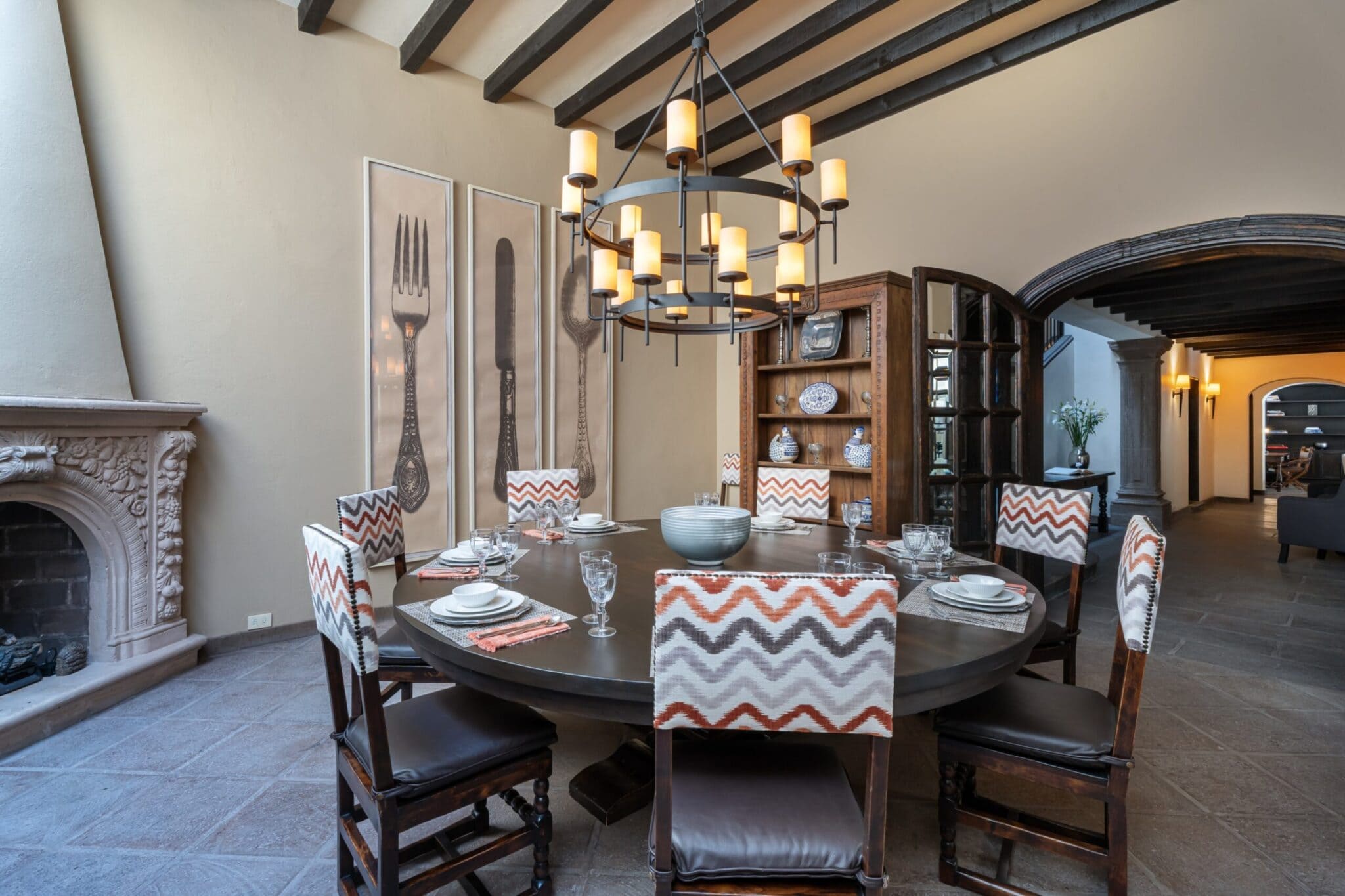 Round dining room with a large wooden table set for a meal, orange zigzag upholstered chairs, and a multi-tier chandelier overhead on a tiled floor, San Miguel de Allende