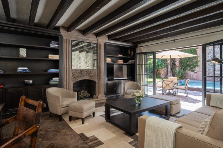 Cozy living room with dark wooden beam ceiling, built-in bookshelves, and a stone fireplace opening to a sunny patio with a pool, San Miguel de Allende