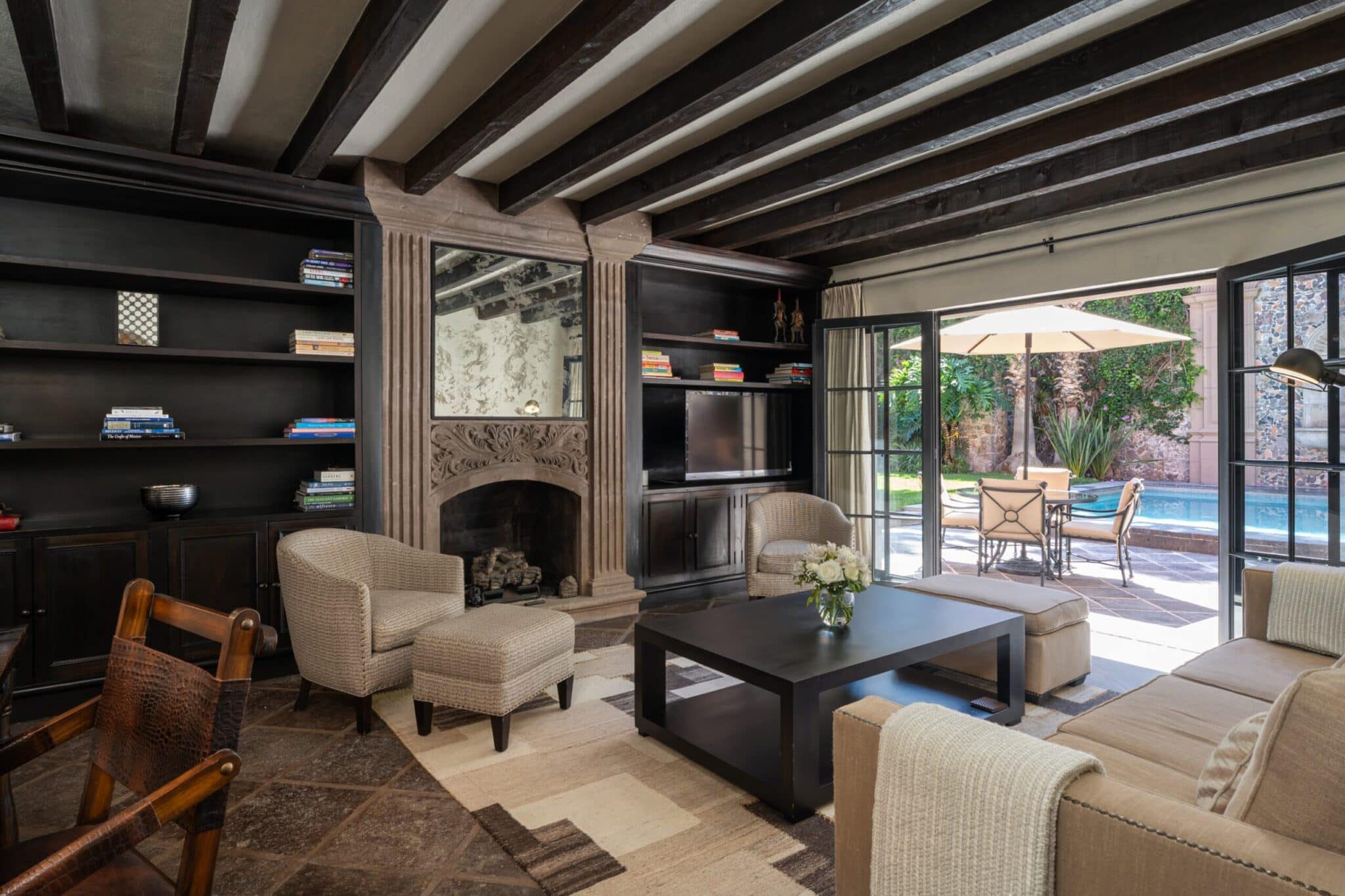 Cozy living room with dark wooden beam ceiling, built-in bookshelves, and a stone fireplace opening to a sunny patio with a pool, San Miguel de Allende