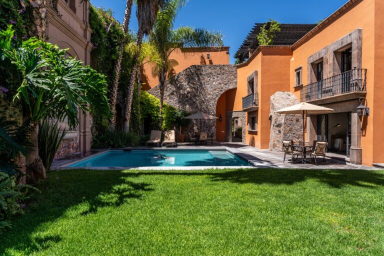 Hotel courtyard with orange stucco walls, a stone arch, palm trees, and a rectangular pool with lounge chairs nearby, San Miguel de Allende