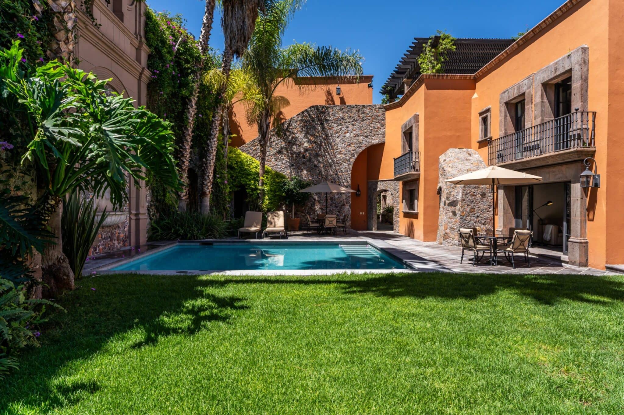 Hotel courtyard with orange stucco walls, a stone arch, palm trees, and a rectangular pool with lounge chairs nearby, San Miguel de Allende