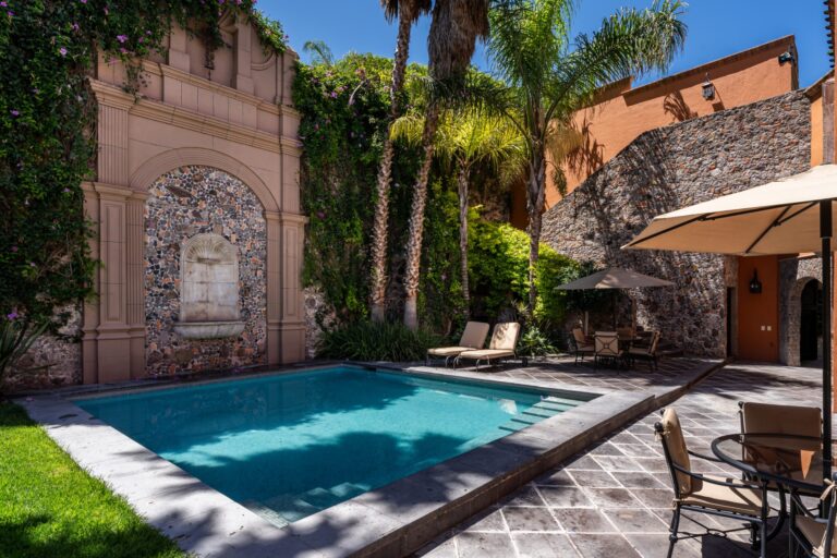 Poolside courtyard with stone walls, palm trees, and beige lounge chairs under umbrellas in bright sun, San Miguel de Allende