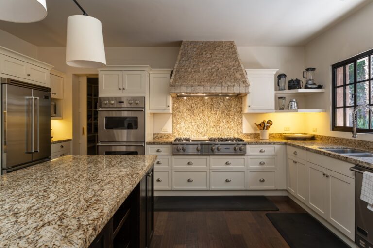 Bright kitchen with granite countertops, white cabinets, and stainless steel double ovens beneath a large stone range hood over a gas cooktop, San Miguel de Allende