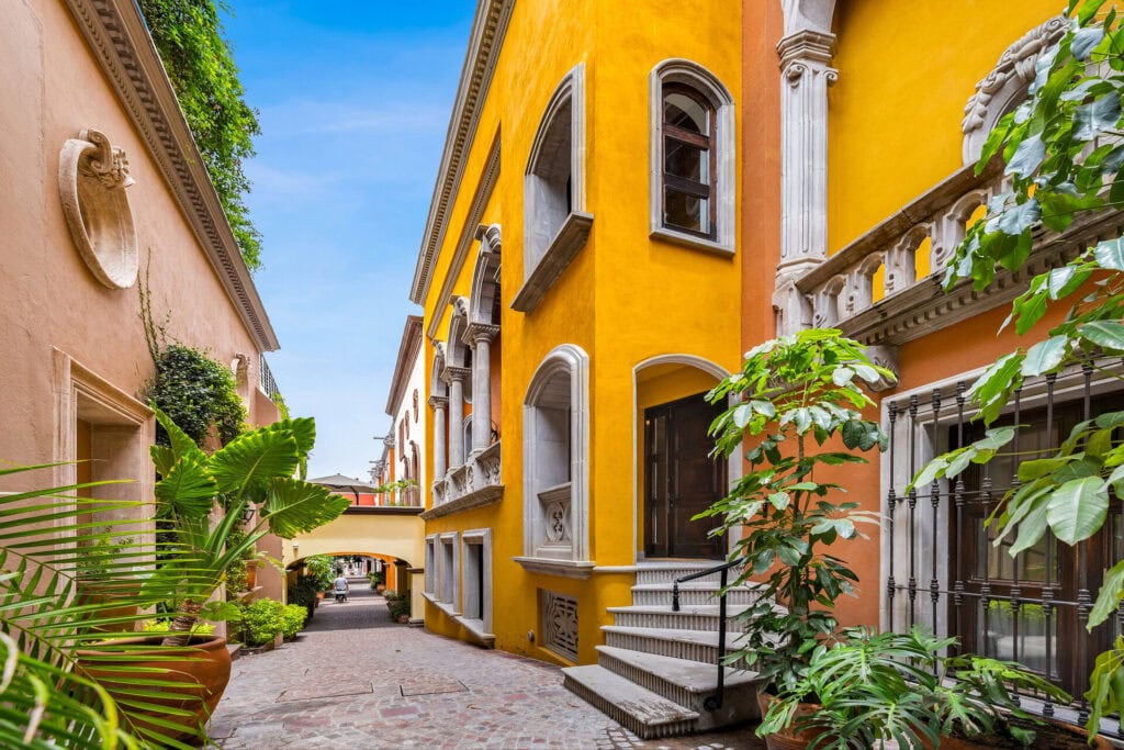 Narrow alley between peach and bright yellow buildings, with arched windows and lush green plants along the walls under a clear blue sky
