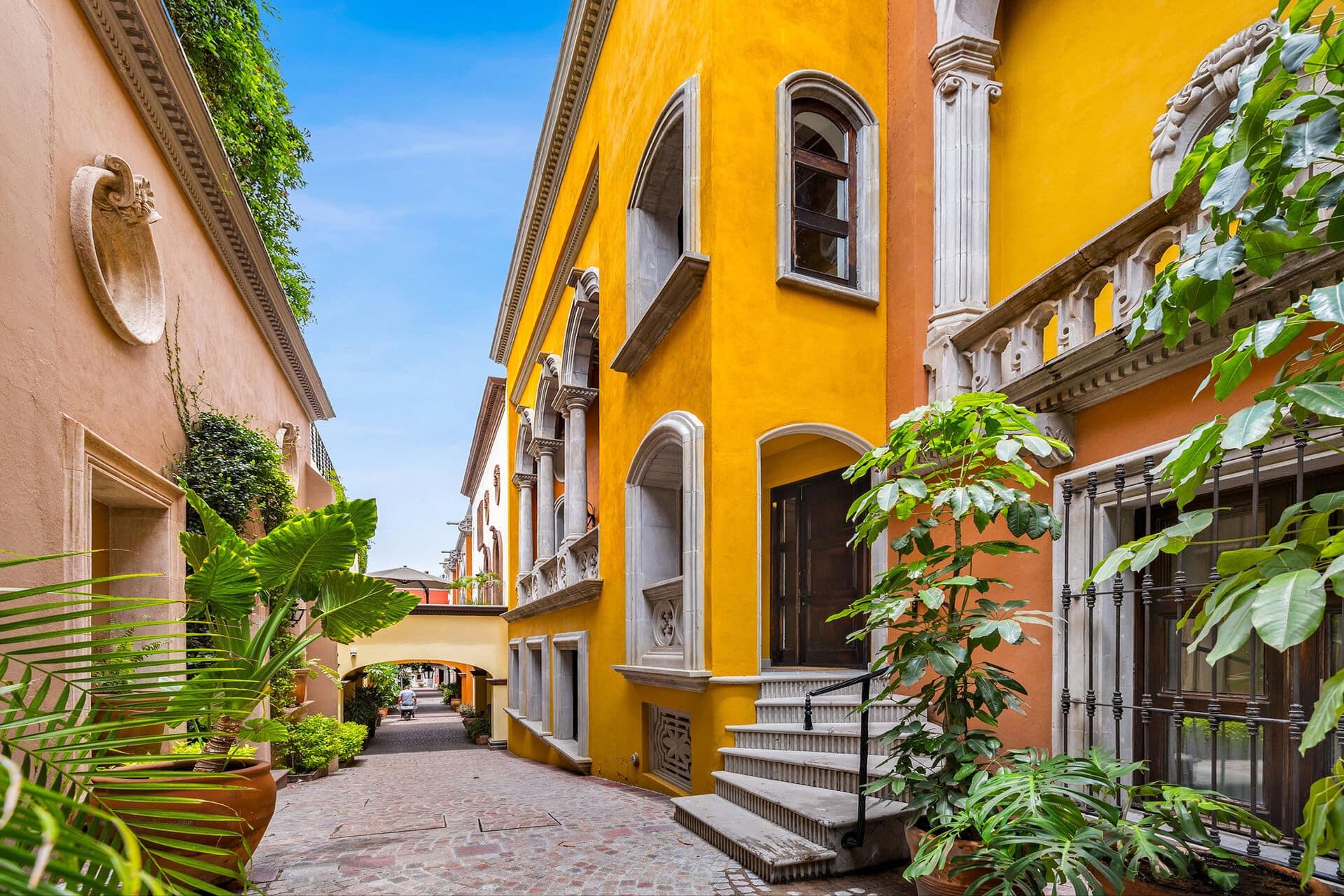 Narrow alley between peach and bright yellow buildings, with arched windows and lush green plants along the walls under a clear blue sky