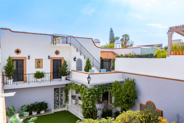 White Mediterranean-style villa courtyard with a stairway, black railing, arched doors, and potted greenery on a sunny day.