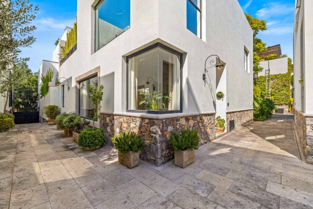 Modern white building corner with large angular windows, stone base, and potted greenery in a sunlit courtyard.