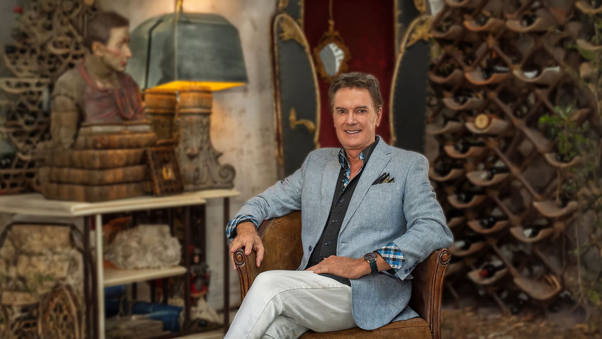 Smiling man in a light blue blazer seated in a wine cellar with bottles on wooden racks behind him.