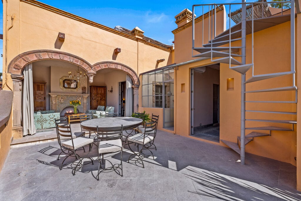 Courtyard with warm yellow walls, arched stone doorway, and an outdoor seating area around a fireplace with sunflowers in a vase.