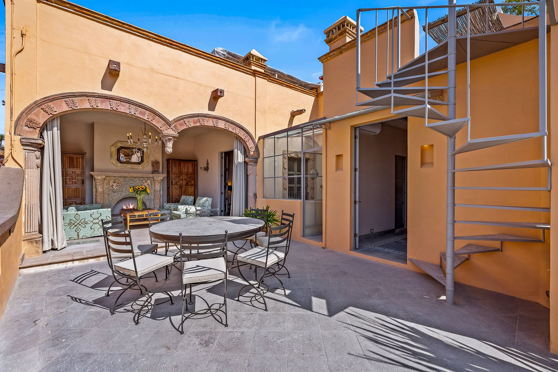 Courtyard with warm yellow walls, arched stone doorway, and an outdoor seating area around a fireplace with sunflowers in a vase.