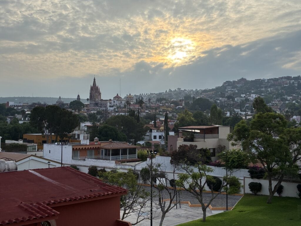 Hillside cityscape at sunset with a tall church spire and red-tiled rooftops in the foreground under a cloudy sky
