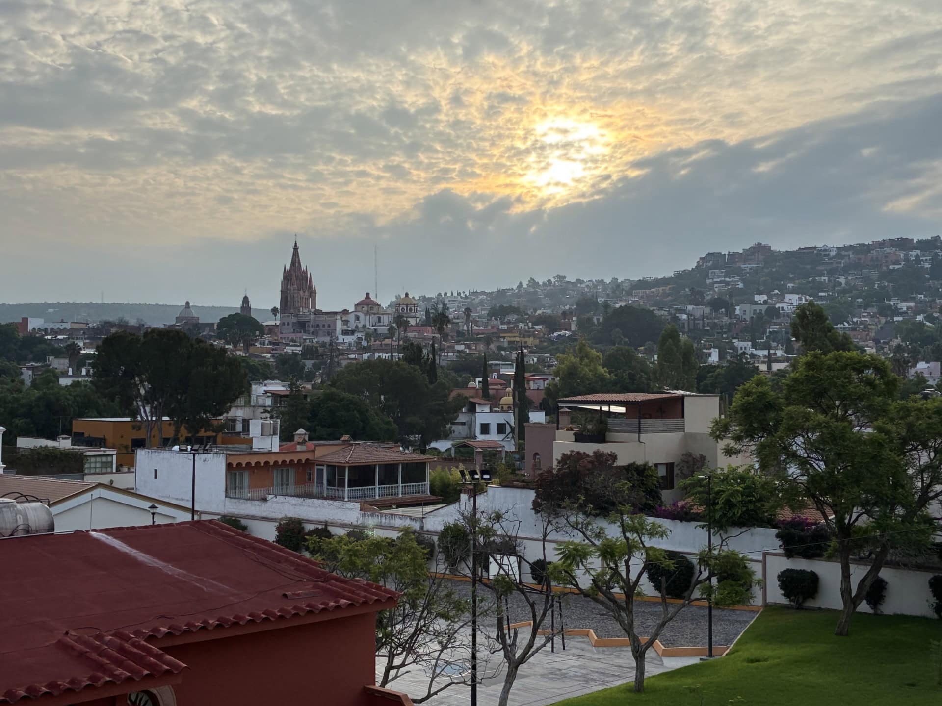 Hillside cityscape at sunset with a tall church spire and red-tiled rooftops in the foreground under a cloudy sky