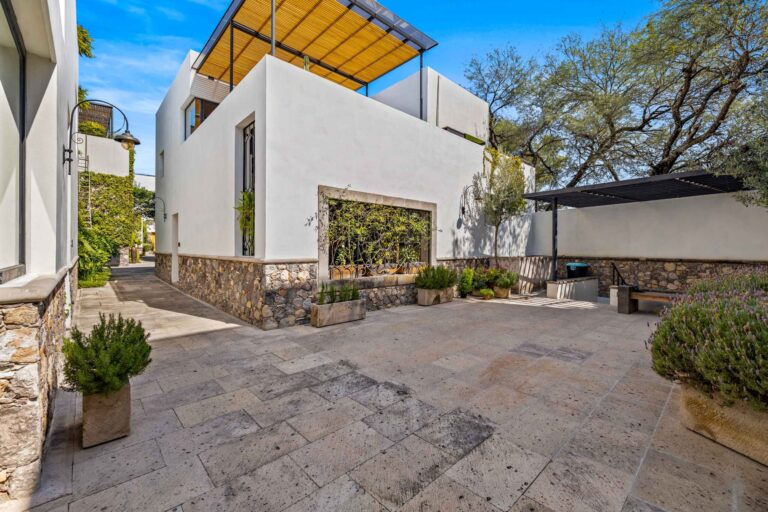 White modern building with stone base, courtyard, and potted plants under a wooden pergola against a blue sky.