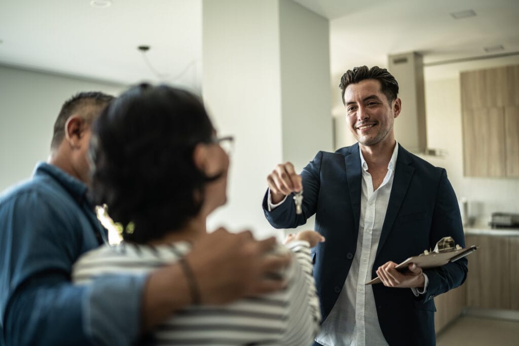 Smiling man in a dark suit hands a key to a group of colleagues in a bright office.