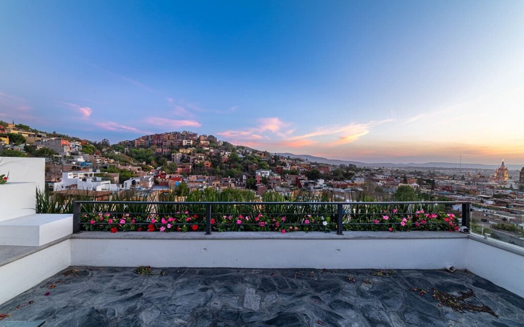 Rooftop terrace with a flower-filled planter and black railing overlooking a hillside city at sunset.