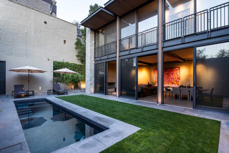 Modern house with glass walls, outdoor seating, and a reflective rectangular poolyard at dusk/early evening