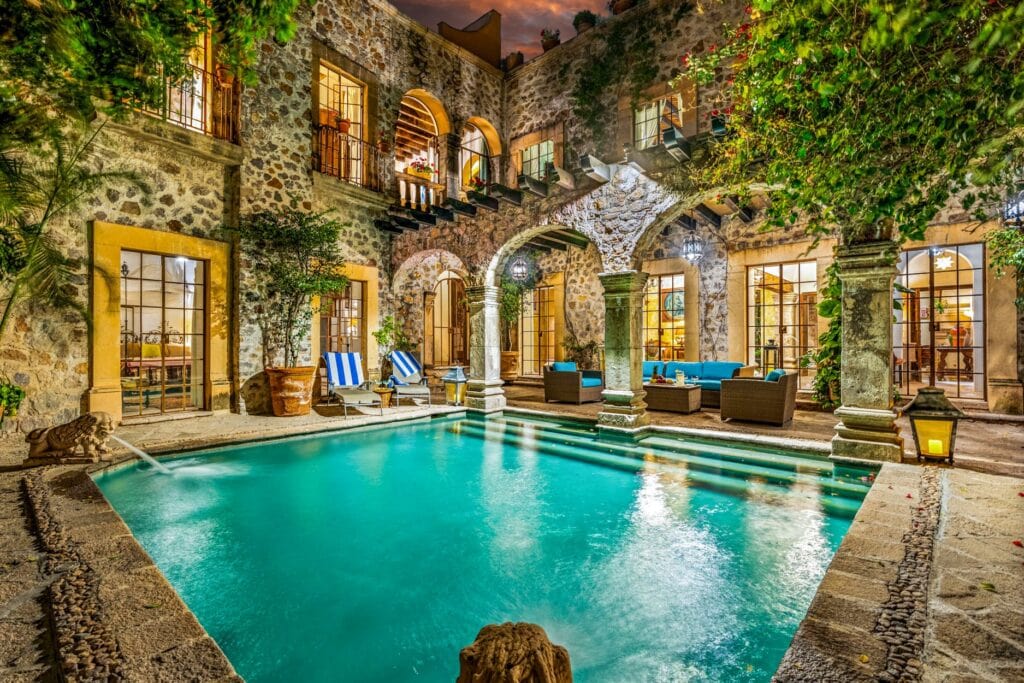 Nighttime stone courtyard with arches, columns, and a turquoise pool, surrounded by greenery and outdoor seating under warm lights.