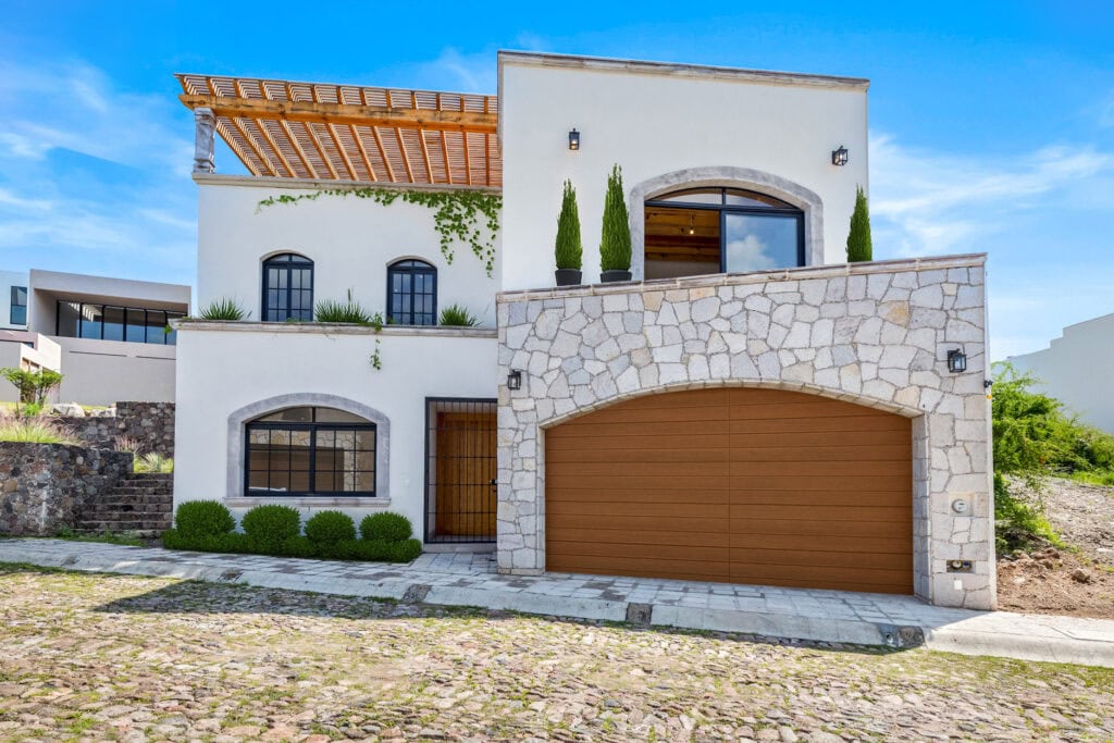 Two-story Mediterranean-style house with a stone arch, tall potted evergreens, and a brown garage door under a blue sky.