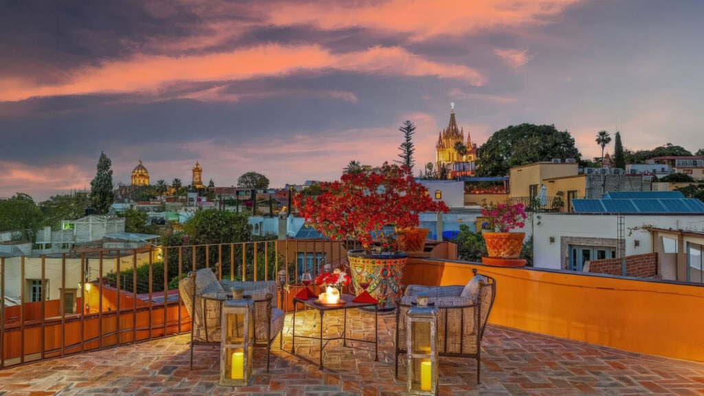 Rooftop terrace at sunset with red flowering plants, candles, and a distant city skyline with church spires.