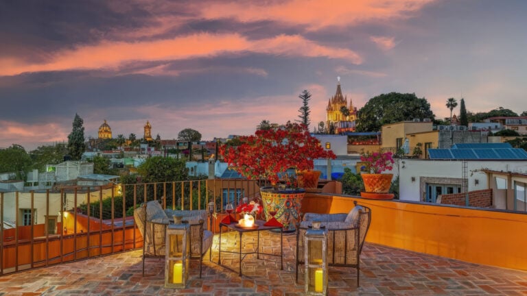 Rooftop terrace at sunset with red flowering plants, candles, and a distant city skyline with church spires.