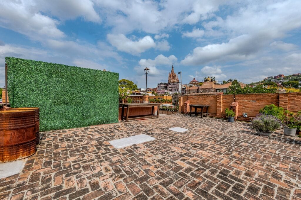 Rooftop terrace with a tall green hedge wall on the left, brick pavement, benches, and a distant city skyline with a church spire under a blue sky with clouds for context.
