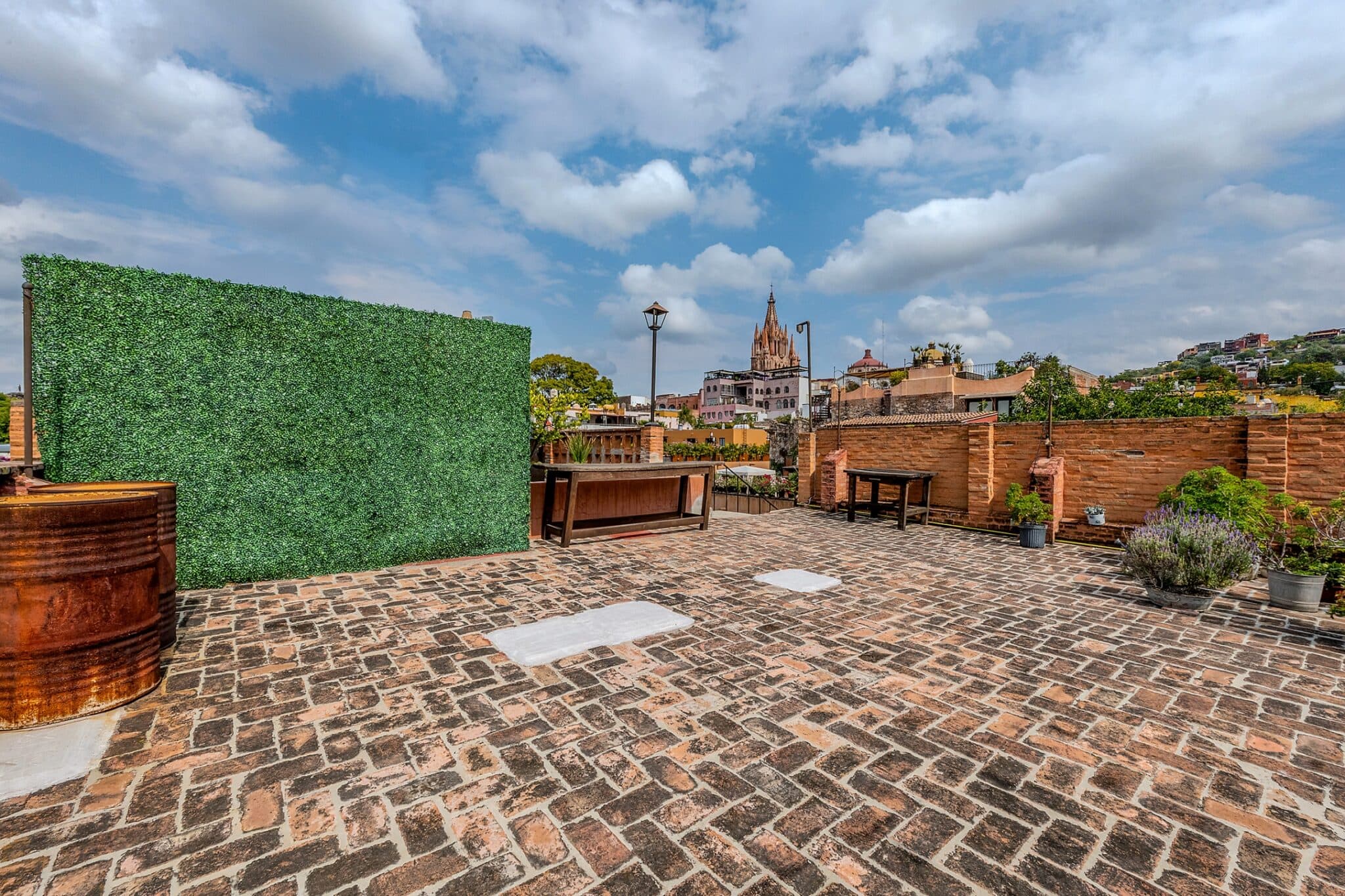 Rooftop terrace with a tall green hedge wall on the left, brick pavement, benches, and a distant city skyline with a church spire under a blue sky with clouds for context.