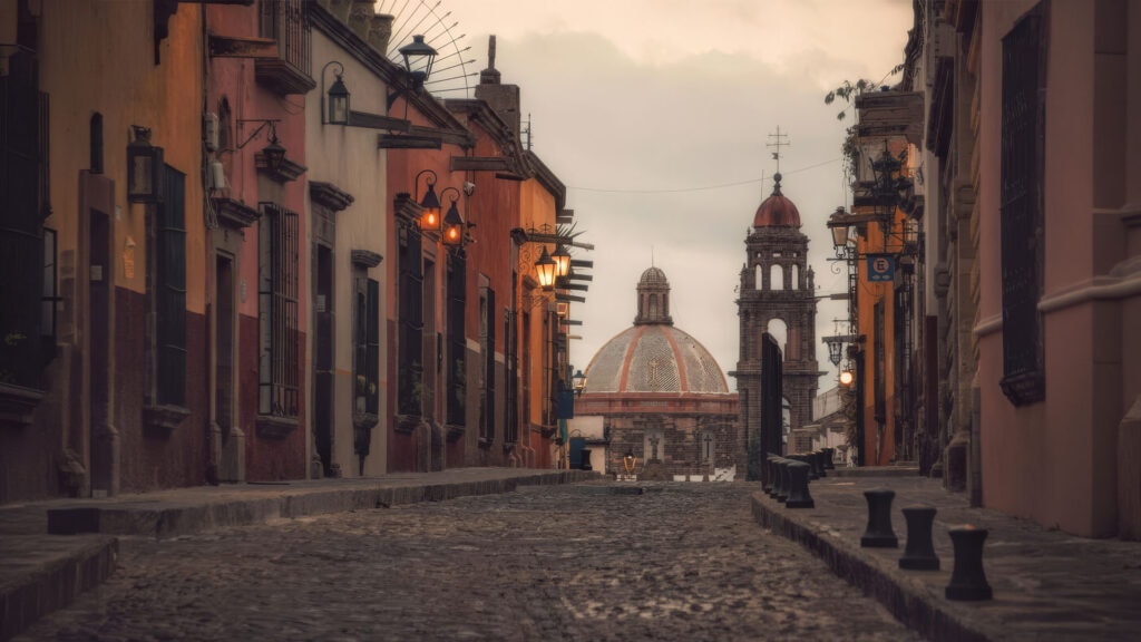 Cobblestone street in a colorful colonial town, with wrought-iron lamps and a distant domed church at dusk.