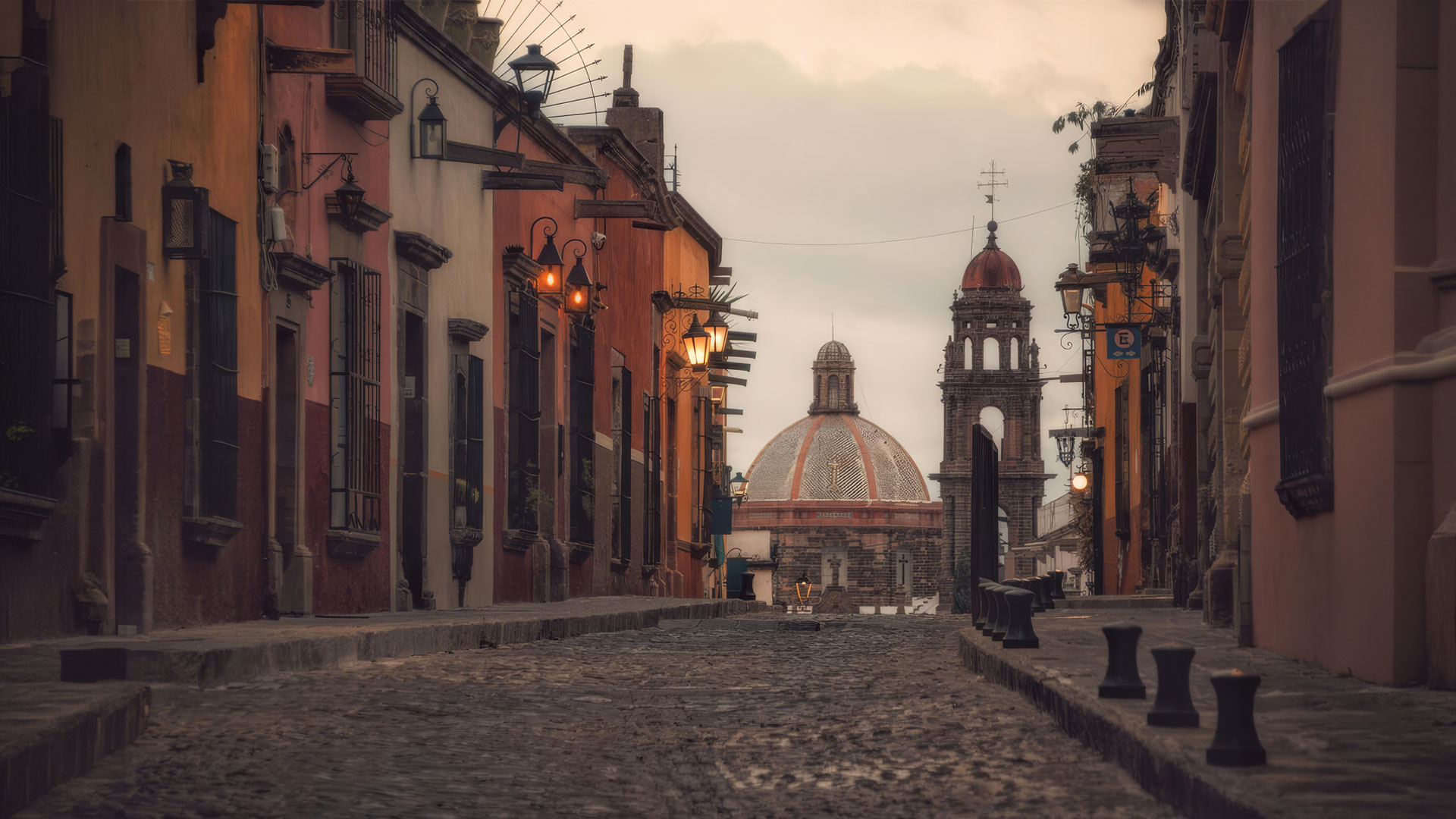 Cobblestone street in a colorful colonial town, with wrought-iron lamps and a distant domed church at dusk.