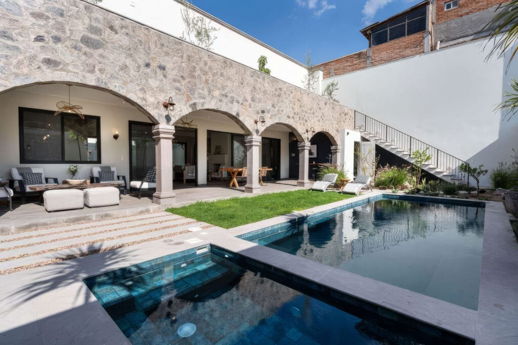 Open-air patio along a stone-walled building with arched doorways, lounge seating, and a narrow rectangular pool reflecting the blue sky.