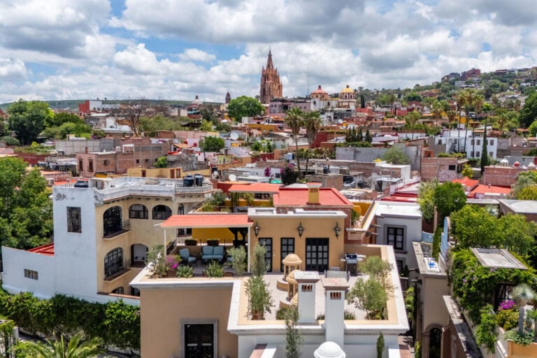 Panoramic view of a colorful Mexican town with a tall cathedral spire, domed churches, and clustered houses under a cloudy sky.