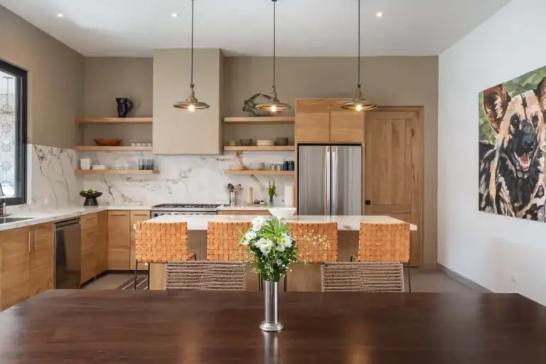 Modern kitchen with marble backsplash, warm wooden cabinetry, an island with seating, and triple pendant lights; foreground shows a dark dining table with a vase of white flowers, San Miguel de Allende