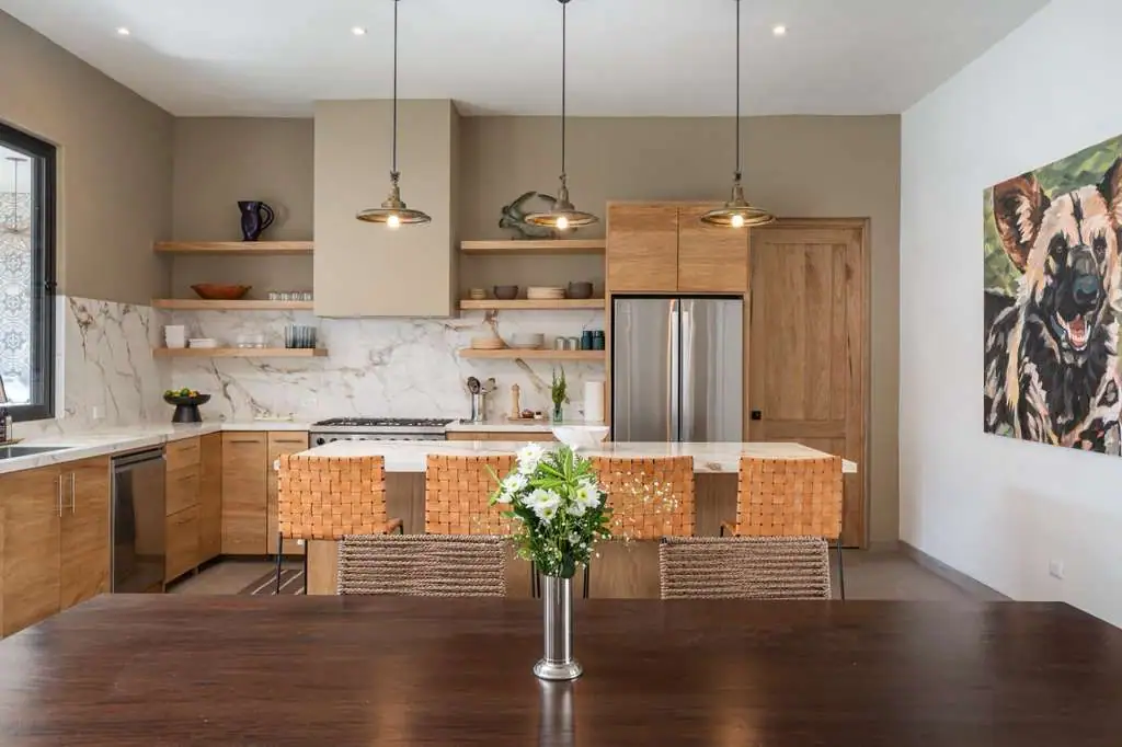 Modern kitchen with marble backsplash, warm wooden cabinetry, an island with seating, and triple pendant lights; foreground shows a dark dining table with a vase of white flowers, San Miguel de Allende
