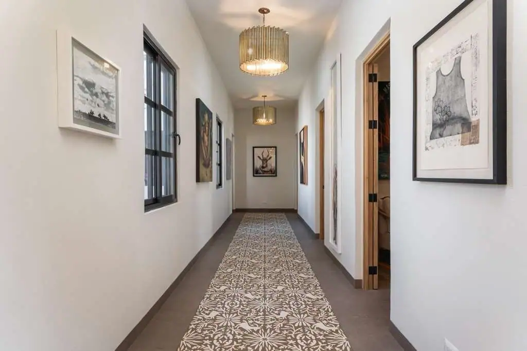 A bright hallway with white walls, framed artworks, and a patterned runner rug leading to a door at the end, two ceiling pendant lights visible, San Miguel de Allende